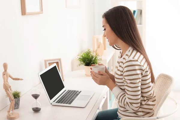 Jovem mulher bebendo café enquanto trabalhava com laptop moderno à mesa — Fotografia de Stock
