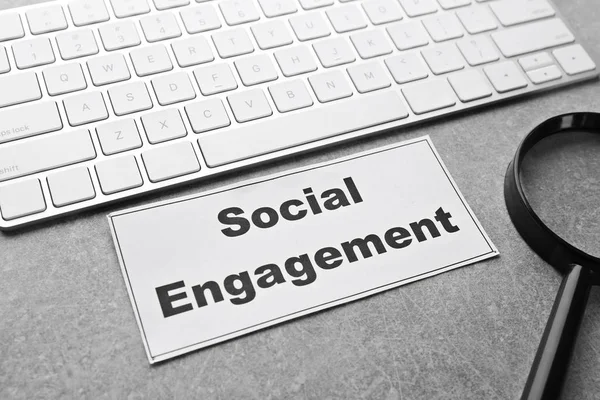 Paper with text SOCIAL ENGAGEMENT near keyboard and magnifier on office table