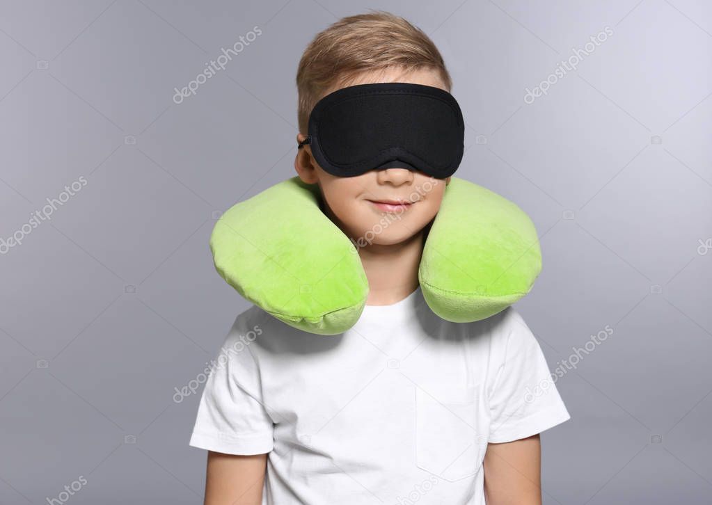 Cute boy with travel pillow and sleep mask on grey background