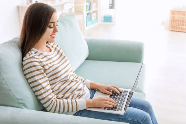 Giovane donna con computer portatile moderno seduta sul divano a casa — Foto Stock