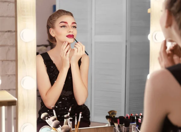 Joven hermosa mujer aplicando maquillaje cerca del espejo en la habitación —  Fotos de Stock