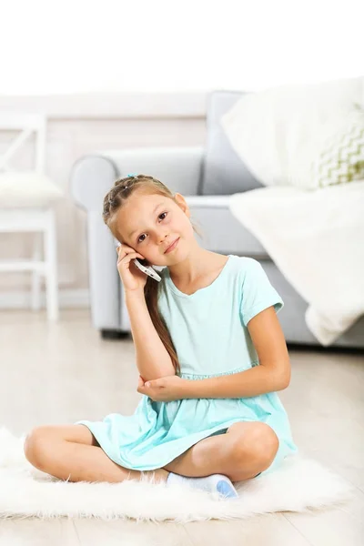 Niña linda con teléfono móvil sentado en la alfombra, en el fondo interior de casa —  Fotos de Stock