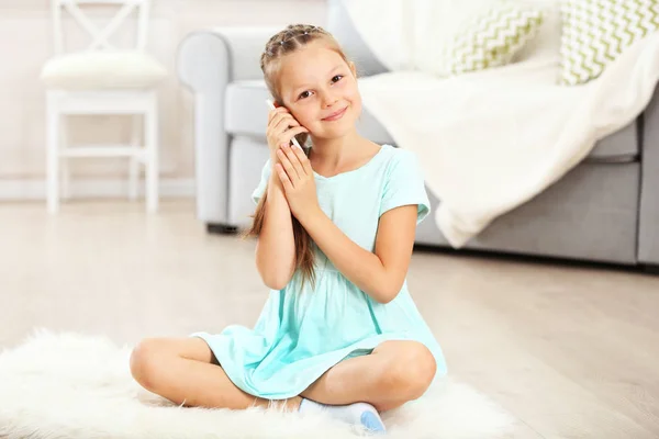 Lilla söta tjejen med mobiltelefon sitter på mattan, på hem interiör bakgrund — Stockfoto
