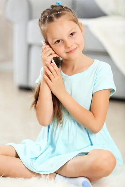 Schattig meisje met mobiele telefoon zittend op het tapijt, op home interieur achtergrond — Stockfoto