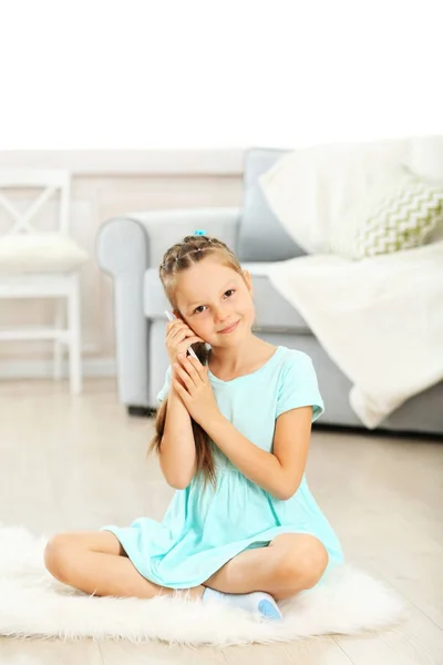 Niña linda con teléfono móvil sentado en la alfombra, en el fondo interior de casa —  Fotos de Stock