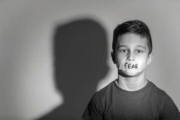 Sad little boy with taped mouth and word "Fear" on grey background, black and white effect