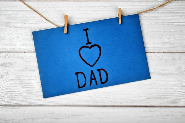Card with phrase I LOVE DAD for Father\'s Day hanging on string against wooden background