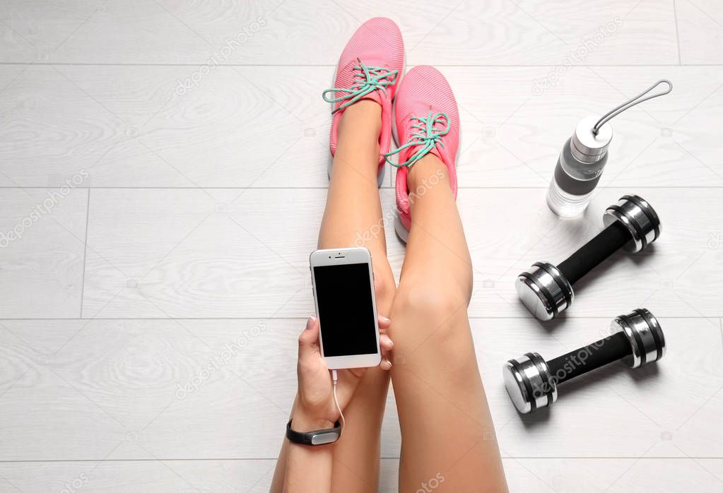 Young woman with mobile phone sitting near dumbbells on floor, flat lay. Ready for gym workout