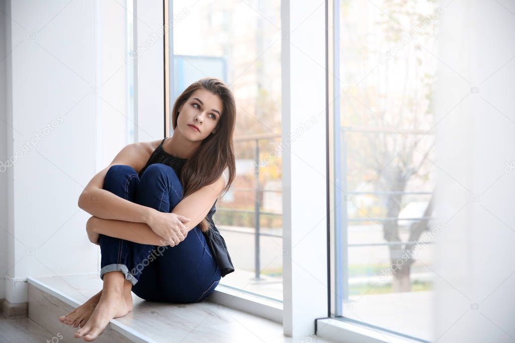 Depressed young woman after breakup sitting on window sill indoors