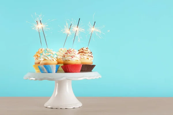 Birthday cupcakes with sparklers on dessert stand against color background