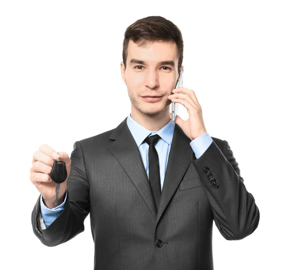 Handsome car salesman with key talking on mobile phone against white background — Stock Photo, Image