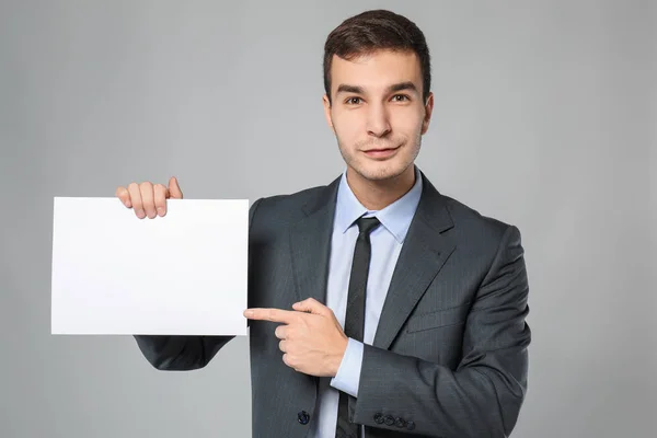 Handsome businessman with blank sheet of paper for advertising on grey background — Stock Photo, Image