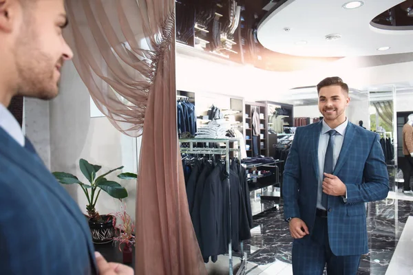 Handsome young man wearing suit looking in mirror at store — Stock Photo, Image