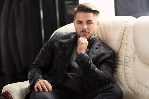 Hombre joven con ropa elegante sentado en el sofá en la tienda de ropa masculina —  Fotos de Stock