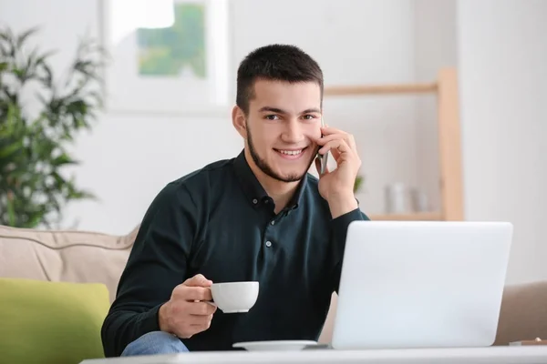 Young man using modern laptop and talking on mobile phone at home — Stock Photo, Image