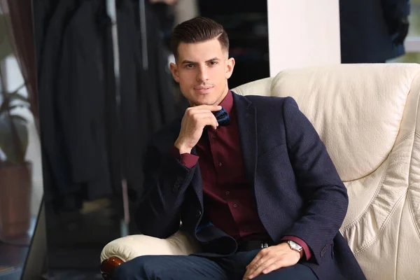 Jovem de terno elegante sentado no sofá na loja de roupa masculina — Fotografia de Stock