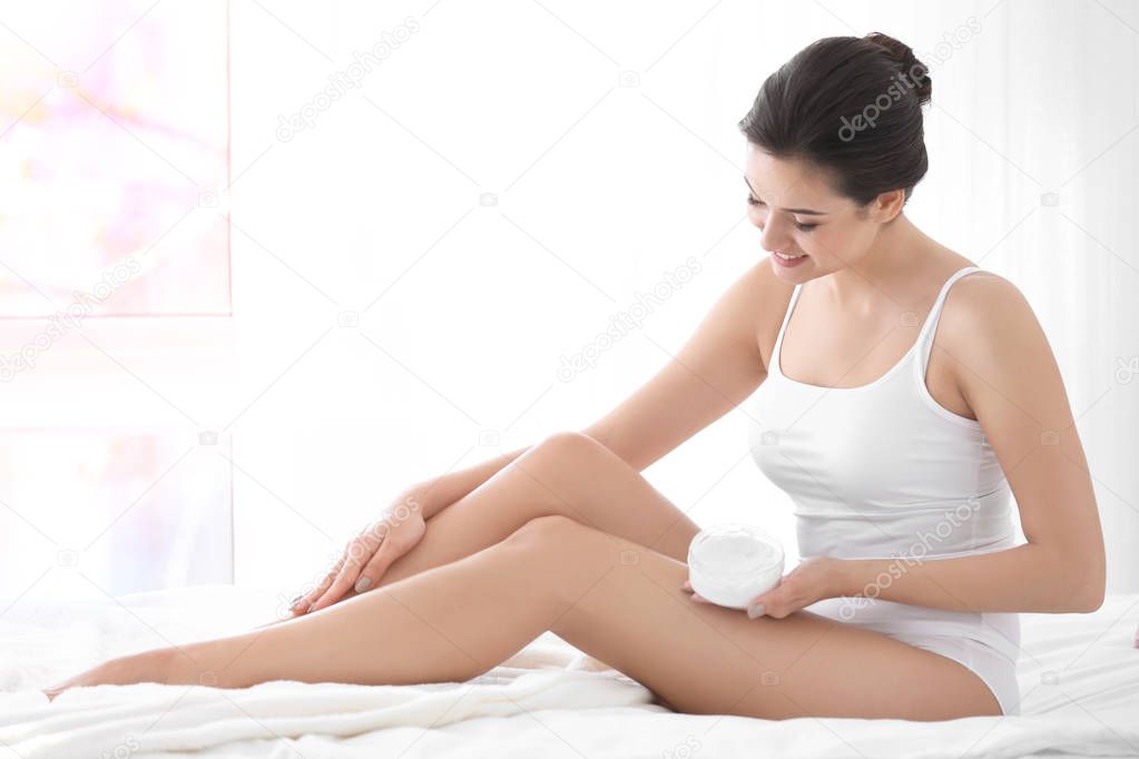 Young woman applying body cream on her leg in bedroom