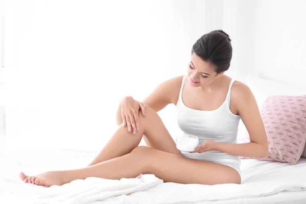 Young woman applying body cream on her leg in bedroom — Stock Photo, Image