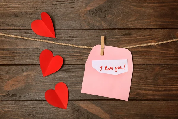 Busta con frase "ti amo" appesa a corda e cuori sullo sfondo di legno — Foto Stock