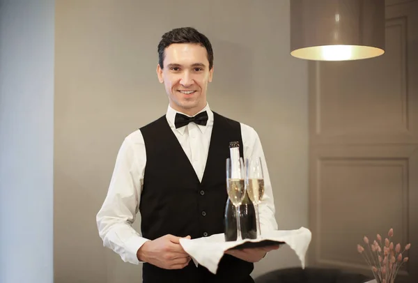 年轻侍者拿着杯子和瓶香槟在室内 — 图库照片