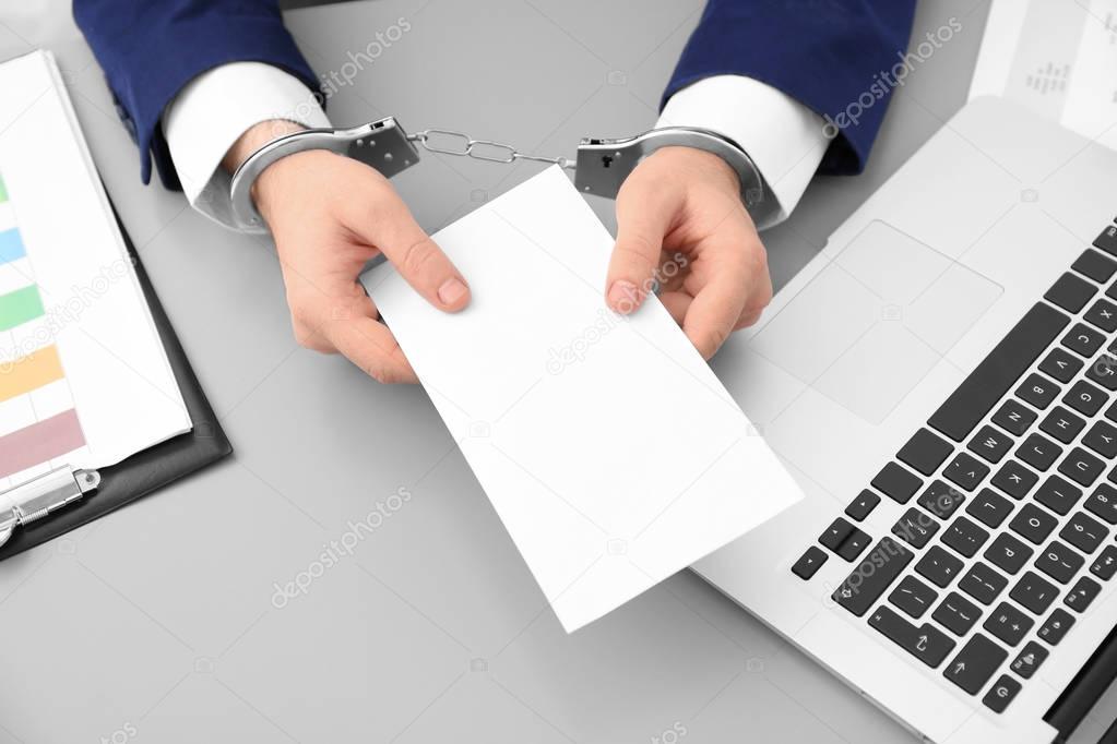 Businessman in handcuffs holding envelope with bribe at table, closeup