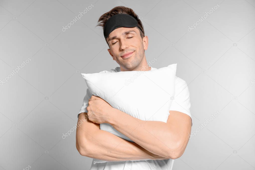 Young man with soft pillow and sleep mask on grey background