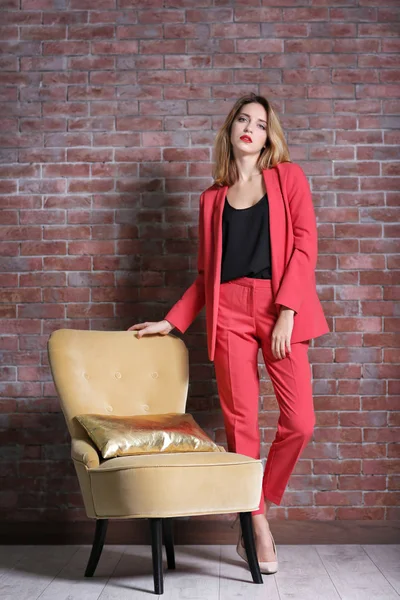 Belle jeune femme élégante debout près du fauteuil contre le mur de briques — Photo