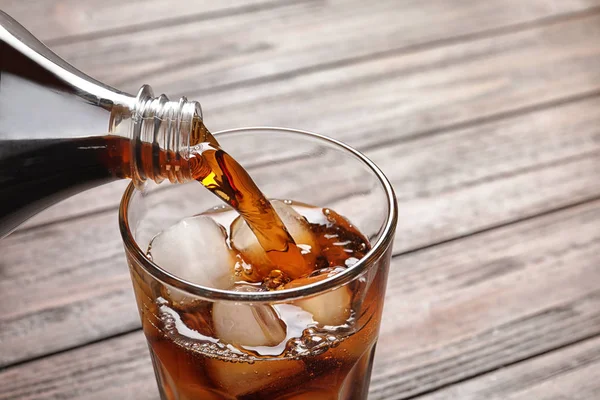 Pouring cola from bottle into glass with ice on wooden background, closeup — Stock Photo, Image