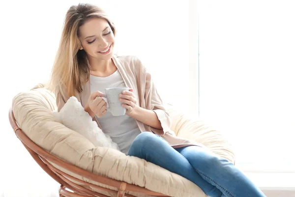 Wanita muda yang bahagia minum teh sambil duduk di kursi di rumah — Stok Foto