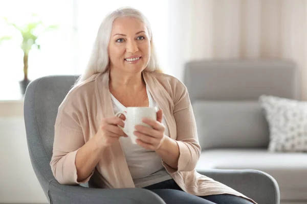 Happy zralá žena pití čaje, zatímco sedí v křesle u vás doma — Stock fotografie