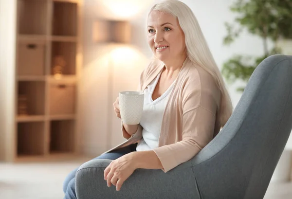 Feliz mujer madura bebiendo té mientras está sentada en el sillón en casa — Foto de Stock
