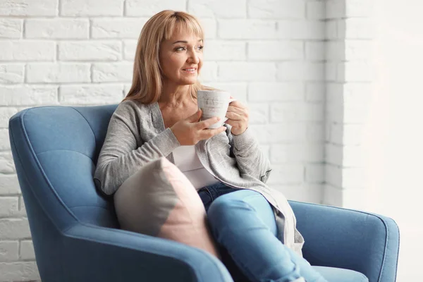 Feliz mujer madura bebiendo té mientras está sentada en el sillón en casa — Foto de Stock