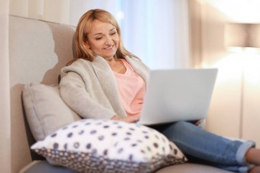 Mutlu olgun kadın evde koltukta otururken dizüstü bilgisayar kullanarak