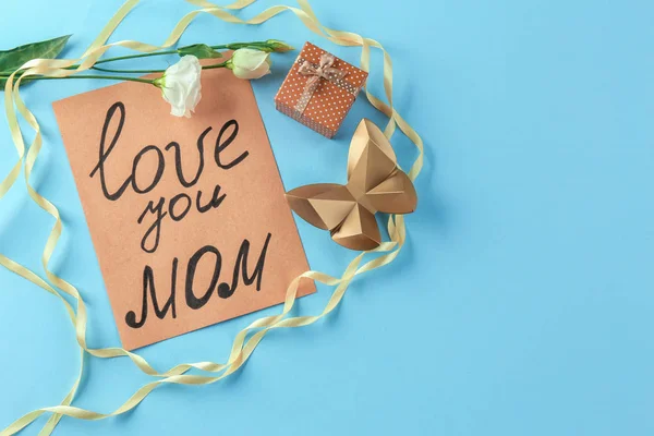 Card with text "LOVE YOU MOM", flower, decorative butterfly and gift box on color background. Greetings for Mother's day — Stock Photo, Image