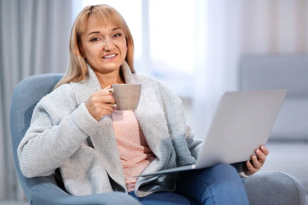 Счастливая зрелая женщина пользуется ноутбуком и пьет чай, сидя дома в кресле — стоковое фото