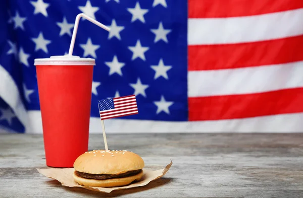 Gustoso hamburger e tazza con bevanda sul tavolo in legno contro la bandiera americana sfocata — Foto Stock