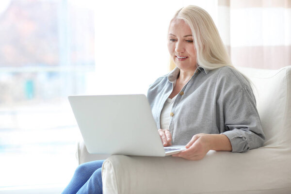 Счастливая зрелая женщина пользуется ноутбуком, сидя дома в кресле
