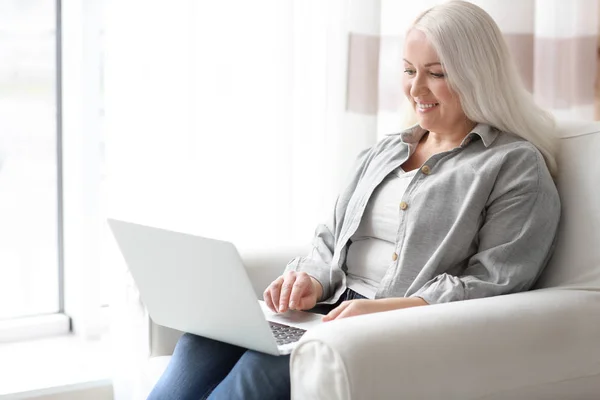 Mulher madura feliz usando laptop enquanto sentado em poltrona em casa — Fotografia de Stock