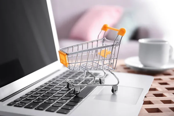 Pequeño carrito de compras y portátil en la mesa, primer plano. Concepto de compras por Internet —  Fotos de Stock