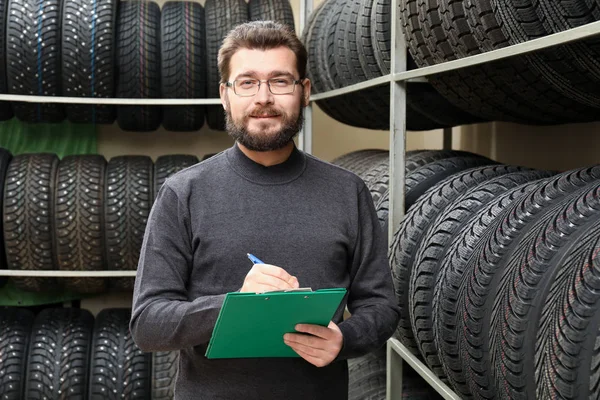 Vendedor con portapapeles cerca de neumáticos en bastidores en la tienda de automóviles — Foto de Stock