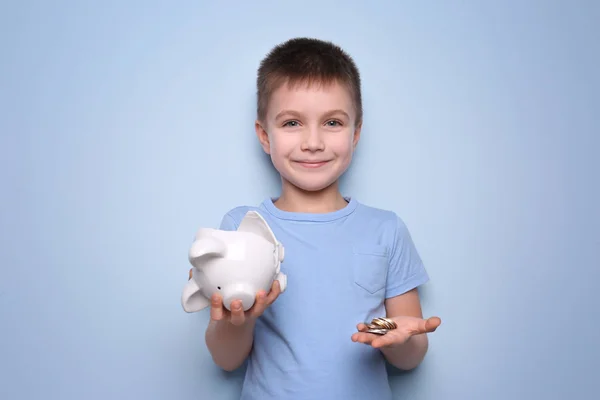 Cute boy holding broken piggy bank and coins on color background — Stock Photo, Image