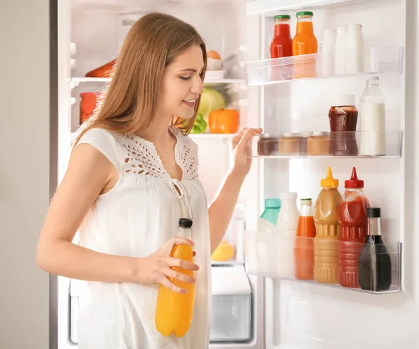 Jonge vrouw met fles drank staande in de buurt van de koelkast — Stockfoto