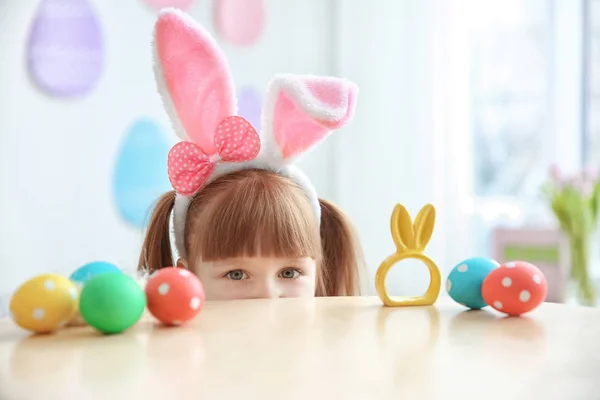 Menina bonito com orelhas de coelho perto da mesa com ovos de Páscoa, dentro de casa — Fotografia de Stock