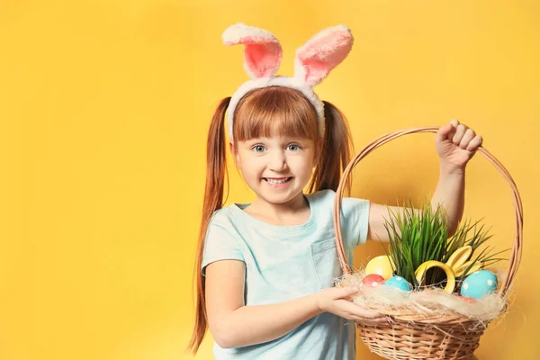 Menina bonito com orelhas de coelho segurando cesta cheia de ovos de Páscoa no fundo de cor — Fotografia de Stock