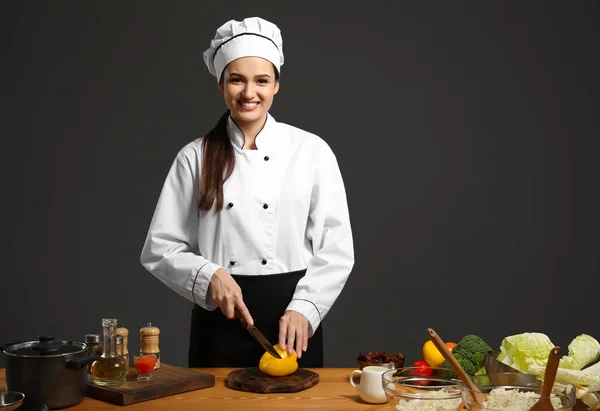 Vackra kvinnliga kocken skär paprika på bordet mot mörk bakgrund — Stockfoto