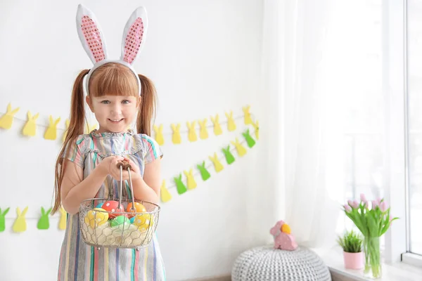 Söt liten flicka med bunny öron holding korg full av färgglada påskägg inomhus — Stockfoto