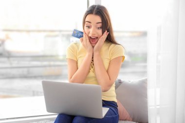 Genç bir kadın evde kredi kartı ve laptop ile online alışveriş yapıyor.