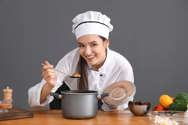Vackra kvinnliga kock provsmakning skålen på bordet mot grå bakgrund — Stockfoto