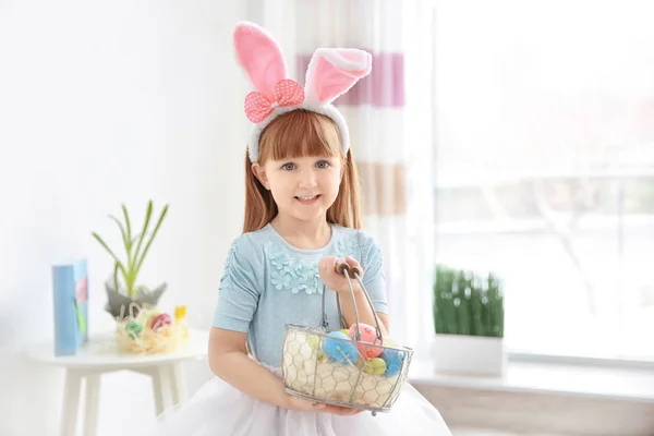 Carino bambina con orecchie di coniglio tenendo cesto pieno di uova di Pasqua colorate al chiuso — Foto Stock