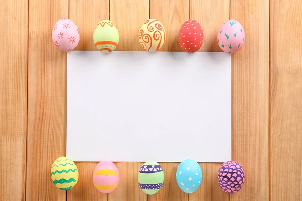 Uova di Pasqua decorate e foglio di carta bianco su sfondo di legno — Foto Stock